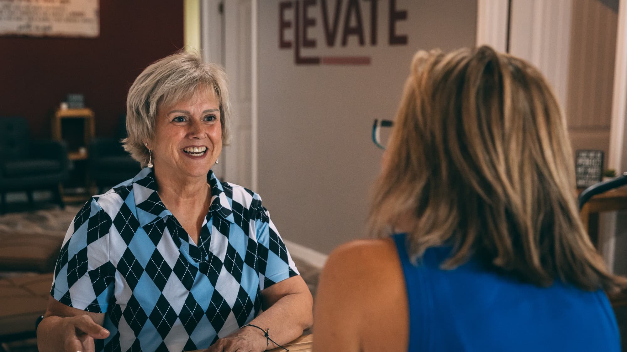 A smiling client in conversation with Elevate founder Jaime Hughes inside the studio, the ELEVATE sign on the wall behind them