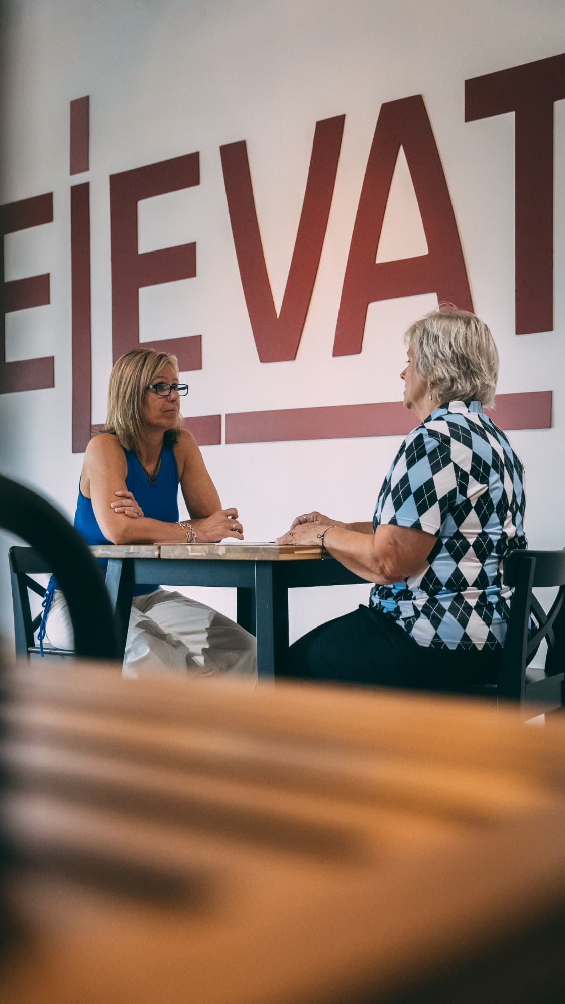 Elevate founder Jaime Hughes talking one-on-one with a client across a table, beneath the large ELEVATE wordmark